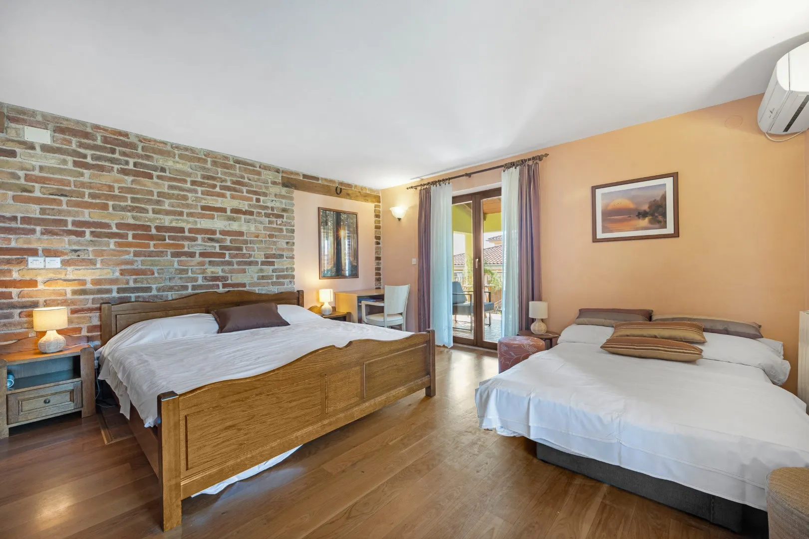 Ein Doppelbett und ein zweites Bett in einem Zimmer mit Sichtziegelwand und Türen zur Terrasse